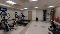 This single-floor fitness room offers a functional and straightforward exercise environment designed to meet essential workout needs. The space features a drop ceiling with recessed fluorescent panel lighting that provides bright, even illumination throughout. The flooring consists of durable vinyl composite tile in a neutral cream and beige pattern, well-suited for high-traffic use and easy maintenance.

The room is equipped with two treadmills, each featuring an integrated monitor screen, positioned side by side along one wall for convenient cardio workouts. A multi-functional cable resistance machine is situated along the adjacent wall, offering a variety of strength training exercises. A small waste bin is neatly placed near the far wall, maintaining the room's tidy appearance.

The walls are finished in a clean, off-white tone, contributing to the bright and open feel of the space. A single interior door provides access to the room, and the overall layout is straightforward and uncluttered, allowing users to move freely between equipment. The space presents a clean, no-frills fitness environment suitable for daily exercise routines.