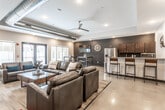 This single-floor communal space showcases a thoughtfully designed open-concept layout that seamlessly blends industrial and contemporary elements. Exposed metallic ductwork runs along the ceiling, complementing the polished concrete floors and contributing to the space's modern, urban character. Recessed lighting and a ceiling fan ensure a well-lit, comfortable atmosphere throughout.

The main lounge area features generously sized dark leather sofas and armchairs arranged around a wooden coffee table, centered on a textured area rug. A wall-mounted flat-screen TV is paired with a soundbar and set against a striking natural stone block accent wall, creating a compelling focal point. Large windows allow natural light to filter in, offering views of the surrounding exterior.

An adjacent dining nook provides high-top tables with bar stools alongside a second wall-mounted TV, making it ideal for casual gatherings or screen viewing. Deep charcoal-painted accent walls in this zone add a bold contrast to the lighter tones elsewhere.

The kitchenette is well-appointed with rich dark wood cabinetry, light quartz countertops, stainless steel appliances including a full-size refrigerator and microwave, and a bar-height peninsula with seating for multiple guests. Decorative touches such as a large clock and a sculptural propeller wall accent add personality to the space. Overall, the design balances functionality with a polished, welcoming ambiance.