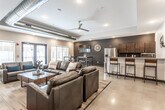 This single-floor communal space showcases a thoughtfully designed open-concept layout that seamlessly blends industrial and contemporary elements. Exposed metallic ductwork runs along the ceiling, complementing the polished concrete floors and contributing to the space's modern, urban character. Recessed lighting and a ceiling fan ensure a well-lit, comfortable atmosphere throughout.

The main lounge area features generously sized dark leather sofas and armchairs arranged around a wooden coffee table, centered on a textured area rug. A wall-mounted flat-screen TV is paired with a soundbar and set against a striking natural stone block accent wall, creating a compelling focal point. Large windows allow natural light to filter in, offering views of the surrounding exterior.

An adjacent dining nook provides high-top tables with bar stools alongside a second wall-mounted TV, making it ideal for casual gatherings or screen viewing. Deep charcoal-painted accent walls in this zone add a bold contrast to the lighter tones elsewhere.

The kitchenette is well-appointed with rich dark wood cabinetry, light quartz countertops, stainless steel appliances including a full-size refrigerator and microwave, and a bar-height peninsula with seating for multiple guests. Decorative touches such as a large clock and a sculptural propeller wall accent add personality to the space. Overall, the design balances functionality with a polished, welcoming ambiance.