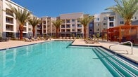 Located at 6504 N 67th Ave in Glendale, Arizona, this outdoor amenity space delivers a resort-style experience designed for relaxation and community living. At its centerpiece is a generously sized swimming pool with crystal-clear blue water, complemented by a separate spa, both framed by wide concrete pool decks lined with lounge chairs ideal for enjoying the abundant Arizona sunshine.

Tall, mature palm trees are strategically placed throughout the courtyard, providing shade and a tropical ambiance that enhances the desert setting. Warm-toned wood pergolas with slatted roofs offer shaded retreats furnished with seating, perfect for socializing or unwinding away from the sun. Additional outdoor dining areas feature tables and chairs set beneath a covered pavilion with recessed lighting, extending the usability of the space into the evening hours.

Thoughtfully curated desert-adapted landscaping borders the walkways and fenced perimeter, incorporating native shrubs, flowering plants, and decorative rock. Wrought-iron fencing with gated access defines the pool area while maintaining open sightlines to the surrounding multi-story residential building, which features bold blue and neutral stucco facades with private balconies overlooking the courtyard. Rocking chairs and casual seating are tucked throughout, creating intimate nooks within this well-appointed outdoor community hub.