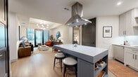 This thoughtfully designed 2-bedroom, 2-bathroom residence on a single floor offers a seamless blend of modern aesthetics and functional living. The open-concept layout connects the kitchen and living room, creating a welcoming, airy atmosphere enhanced by light wood flooring throughout.

The kitchen is a standout feature, centered around a large island with a white quartz countertop, bar seating, and a striking stainless steel range hood overhead. Light gray shaker-style cabinetry, stainless steel appliances, and a clean subway tile backsplash complete the space. The adjacent living room is bright and inviting, with large floor-to-ceiling glass doors opening to a private balcony with outdoor views, and a modern globe chandelier adding an elegant focal point.

The primary bedroom is a comfortable retreat with natural light from a large window and direct access to a spacious walk-in closet outfitted with customizable shelving and a built-in bench. The en-suite bathroom features slate-blue cabinetry, stone tile, a soaking tub, and refined matte black hardware. The second bedroom is equally well-lit with outdoor views. The second bathroom mirrors the design quality with coordinating blue cabinetry and tub-shower combination. Two closets provide ample storage throughout the residence.