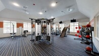 This fitness center at 16755 W Lake Houston Parkway in Houston, Texas, offers a generously sized, single-level workout space designed to accommodate a wide variety of training styles. The interior features vaulted ceilings in select areas, adding a sense of openness and volume throughout the room. A bold orange accent band runs along the upper portion of the walls, contrasting with the neutral white walls and blue-striped commercial carpeting underfoot, lending the space a modern, energetic aesthetic.

Natural light enters through multiple white-framed windows positioned along the perimeter walls, keeping the environment bright and welcoming. Wall-mounted flat-screen televisions are strategically placed throughout the space for entertainment during workouts. Full-length mirrors line portions of the walls, enhancing the sense of space while providing visual feedback for exercisers.

The equipment layout is thoughtfully arranged to maximize usable floor space. A multi-functional cable and functional trainer system serves as a centerpiece, accompanied by a stair climber, ellipticals, treadmills, and a rowing machine for cardiovascular training. The strength area includes a squat rack, free weights, benches, and various selectorized machines. Battle ropes and resistance accessories round out the functional fitness offerings. Access is provided via glass entry doors, and sanitation stations are conveniently mounted on the walls.