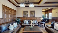 Located at 2000 Villa Dr in Pittsburg, CA, this single-floor space at San Marco Villas offers a sophisticated and warmly appointed interior with a living room and dining room, unified by a cohesive Mediterranean-inspired aesthetic. Throughout the space, rich dark wood wainscoting and wall paneling create a refined backdrop, complemented by warm beige walls and intricately patterned carpeting that runs seamlessly across both rooms.

The living room features a coffered ceiling with dark wood crown molding and beam accents that lend architectural depth and height to the space. Elegant ironwork double doors with ornate scrollwork designs serve as a striking entryway, while leather sofas and a wood coffee table complete the inviting seating arrangement. Framed artwork and a pendant light fixture add to the room's polished character.

The dining room is bathed in natural light streaming through large plantation-shuttered windows framed in rich wood, offering views of the surrounding exterior landscape. A substantial dark wood dining table surrounded by classic chairs anchors the room, while lush potted plants add a fresh, organic element. The tray ceiling with warm ambient lighting further elevates the dining experience, making this a welcoming and detail-rich space throughout.
