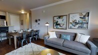 This single-floor residence offers a well-appointed open-concept layout that seamlessly connects the living and dining areas, creating a cohesive and functional living environment. The living area is anchored by a contemporary gray sofa accented with decorative throw pillows, complemented by a plush area rug and a sleek metallic coffee table that adds a touch of glamour. A modern arc floor lamp provides warm ambient lighting, while a pair of large abstract framed artworks adorns the neutral-toned walls, contributing to the space's curated aesthetic. Crown molding runs along the ceiling, adding an elegant architectural detail that enhances the room's refined character. The adjacent dining area features a rustic wood table paired with industrial-style metal chairs, offering a striking contrast that balances the overall design. Dark hardwood flooring flows throughout, grounding the space with warmth and visual continuity. The neutral wall palette allows the furnishings and artwork to take center stage, while decorative wall accents add personality and depth. The open layout promotes a natural flow between entertaining and everyday living spaces, making this residence both inviting and practical.