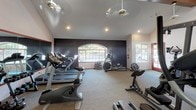 Located at 15095 Via Hondonada in the Rancho Peñasquitos neighborhood of San Diego, California, this single-floor fitness center offers a well-equipped workout space designed for both comfort and functionality. The room features vaulted ceilings with recessed lighting and ceiling fans, ensuring ample air circulation and a bright, open atmosphere. A bold, dark accent wall serves as a striking architectural focal point, contrasting cleanly with the lighter surrounding walls and neutral carpet flooring. Natural light flows in through an arched window and additional side windows, providing views of the surrounding greenery and creating an inviting exercise environment. The space is outfitted with a variety of cardio machines, including treadmills, an elliptical trainer, a stationary bike, and a rowing machine, accommodating a range of workout preferences. Additional amenities include a wall-mounted flat-screen display and a portable floor fan for supplemental airflow. The layout is thoughtfully arranged to allow for comfortable movement between equipment. This fitness center presents a clean, functional, and well-maintained facility suited for daily exercise routines.