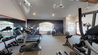 Located at 15095 Via Hondonada in the Rancho Peñasquitos neighborhood of San Diego, California, this single-floor fitness center offers a well-equipped workout space designed for both comfort and functionality. The room features vaulted ceilings with recessed lighting and ceiling fans, ensuring ample air circulation and a bright, open atmosphere. A bold, dark accent wall serves as a striking architectural focal point, contrasting cleanly with the lighter surrounding walls and neutral carpet flooring. Natural light flows in through an arched window and additional side windows, providing views of the surrounding greenery and creating an inviting exercise environment. The space is outfitted with a variety of cardio machines, including treadmills, an elliptical trainer, a stationary bike, and a rowing machine, accommodating a range of workout preferences. Additional amenities include a wall-mounted flat-screen display and a portable floor fan for supplemental airflow. The layout is thoughtfully arranged to allow for comfortable movement between equipment. This fitness center presents a clean, functional, and well-maintained facility suited for daily exercise routines.