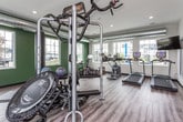 This fitness center is a thoughtfully designed, single-floor facility that offers a comprehensive workout environment. The space is defined by wood-look flooring throughout, recessed lighting, and exposed industrial-style ductwork that runs along the ceiling, lending the interior a modern, functional aesthetic. Large, multi-pane windows line multiple walls, flooding the space with natural light and providing views of the surrounding exterior.

The facility is organized to accommodate a wide range of exercise needs. A dedicated cardio zone features treadmills, ellipticals, a stationary bike, and rowing machines arranged for comfortable use. Wall-mounted televisions are positioned for visibility from cardio equipment. A strength training area includes a multi-station cable machine, a functional trainer, and additional resistance equipment. A free weight section offers a full dumbbell rack alongside an adjustable bench and rubber floor mats for safety.

Accent walls painted in deep forest green provide visual contrast against the lighter neutral tones of the remaining walls. Full-length mirrors are strategically placed to enhance the sense of openness and assist users during exercise. The space also includes stability balls and other accessory equipment, rounding out a well-stocked, versatile fitness facility.
