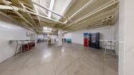 This single-floor utility space offers a generously open layout with an industrial aesthetic defined by its key architectural elements. The polished concrete floors extend throughout, providing a durable and low-maintenance foundation. Overhead, an exposed ceiling reveals a network of pipes, conduits, and structural beams painted white, contributing to the space's utilitarian character while maintaining a clean appearance.

The open floor plan is anchored by several structural concrete columns, which divide the space into functional zones without fully enclosing them. A dedicated laundry area houses multiple washing machines and dryers, accompanied by a folding table for convenience. Nearby, a wall-mounted mailbox bank serves as a centralized mail collection point. Vending machines—including a Pepsi unit and a snack machine—are positioned along one wall, offering easy access to refreshments.

Additional furnishings include a small seating area with teal chairs, a bar-height table with stools, and a bench. Bicycle storage is available along one wall. Fluorescent lighting supplements the limited natural light entering through small windows. A clearly marked exit door ensures safe egress. Overall, the space functions as a practical, multi-purpose amenity area with an open and utilitarian ambiance.
