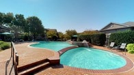 This single-story amenity complex showcases a thoughtfully designed outdoor living environment centered around a generously sized resort-style swimming pool. The pool features elegant water fountains and an integrated spa with brick coping, creating a standout focal point within the space. Multiple lounge chairs line the pool deck, which is finished in herringbone-patterned brick pavers that lend a warm, classic aesthetic throughout the outdoor areas.

A covered arcade runs along the building's exterior, supported by distinctive textured columns with arched detailing. This shaded corridor is furnished with wicker seating and navy blue cushions, offering a comfortable retreat with direct views of the pool and surrounding greenery. Large bay windows and floor-to-ceiling glass panels along the building facade flood adjacent interior spaces with natural light and frame views of the outdoor amenities.

A separate pergola-covered patio provides additional outdoor seating with bistro-style tables and chairs, ideal for relaxing or socializing beneath dappled sunlight filtered through the dark wood lattice overhead. The space is framed by mature trees, manicured hedges, and well-maintained landscaping that enhances the sense of privacy and tranquility. The overall design reflects a Mediterranean-influenced architectural style, combining stucco exteriors, tile rooflines, and brick accents to create a cohesive and inviting communal environment.