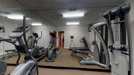 Located at 5811 Spring Run Dr in Columbus, Ohio, this single-floor fitness space is purpose-built for exercise and physical wellness. The dedicated workout room is the centerpiece of the facility, outfitted with a well-rounded selection of fitness equipment including multiple elliptical machines, a treadmill, a weight bench, and a multi-function cable strength training machine. The room is finished with neutral gray walls and carpeted flooring suited for gym use. Full-length mirrored walls span the length of the space, creating a sense of openness and allowing users to monitor their form during workouts. Overhead fluorescent lighting provides bright, even illumination throughout the room. The mirrored walls enhance the feeling of spaciousness, making the workout area feel larger and more expansive than its footprint suggests. A doorway visible through the mirrors connects to adjacent areas of the building, offering convenient access throughout the facility. The overall ambiance is clean, functional, and focused, providing an environment well-suited for a productive fitness routine.