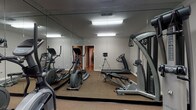 Located at 5811 Spring Run Dr in Columbus, Ohio, this single-floor fitness space is purpose-built for exercise and physical wellness. The dedicated workout room is the centerpiece of the facility, outfitted with a well-rounded selection of fitness equipment including multiple elliptical machines, a treadmill, a weight bench, and a multi-function cable strength training machine. The room is finished with neutral gray walls and carpeted flooring suited for gym use. Full-length mirrored walls span the length of the space, creating a sense of openness and allowing users to monitor their form during workouts. Overhead fluorescent lighting provides bright, even illumination throughout the room. The mirrored walls enhance the feeling of spaciousness, making the workout area feel larger and more expansive than its footprint suggests. A doorway visible through the mirrors connects to adjacent areas of the building, offering convenient access throughout the facility. The overall ambiance is clean, functional, and focused, providing an environment well-suited for a productive fitness routine.