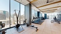 Located in the heart of Salt Lake City at 201 E 300 S, this well-appointed fitness center offers a spacious, single-floor layout designed to accommodate a comprehensive workout experience. The facility is defined by dramatic floor-to-ceiling windows that line the perimeter, flooding the space with abundant natural light and framing sweeping panoramic views of the downtown Salt Lake City skyline and the surrounding mountains.

The interior design blends a modern aesthetic with functional purpose. Muted blue accent walls contrast cleanly with light tile flooring laid in a large-format grid pattern, while wood-beam detailing and recessed lighting in the ceiling add warmth and architectural character. Ceiling fans are integrated throughout, contributing to a comfortable environment during workouts.

The gym is comprehensively equipped with a dedicated cardio zone featuring multiple treadmills, elliptical trainers, rowing machines, stair climbers, and a StairMaster. A strength training area houses a full rack of dumbbells, adjustable benches, cable machines, and a multi-function weight station. Large mirrors are strategically placed to enhance the sense of openness and assist with form. The space also includes a hydration station and ancillary amenities near the entry. The overall ambiance is bright, energizing, and professionally designed for serious fitness use.