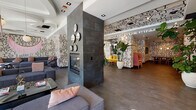 Located on Chandler Boulevard in Los Angeles, this single-floor space makes an immediate impression with its bold, floor-to-ceiling black-and-white graphic mural artwork that envelops nearly every wall and extends across select ceiling surfaces. The continuous abstract line-art motif gives the entire space a cohesive, artistic identity while creating an energetic visual backdrop throughout.

The open layout flows seamlessly across multiple lounge and seating zones. A central dark-slate-tiled fireplace column anchors the main area, flanked by clusters of plush grey sofas accented with blush pink cushions. A striking pink fringe chandelier with gold accents adds a glamorous, eclectic touch overhead, complemented by sculptural coffered ceiling details in one lounge section.

A vibrant alcove features a bold mustard-yellow sectional sofa set beneath wall-mounted shelving displaying curated objects, lit by globe pendant lights. Lush tropical plants in large white ceramic planters are positioned throughout, introducing natural warmth and organic contrast to the graphic interiors.

Additional seating areas include pink high-backed booth-style chairs and gold-framed accent furniture. Steel-framed glass partitions delineate a separate interior room, visible from the main corridor. Polished large-format tile flooring runs throughout, and full-height glass entry doors invite ample natural light from the exterior.
