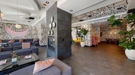 Located on Chandler Boulevard in Los Angeles, this single-floor space makes an immediate impression with its bold, floor-to-ceiling black-and-white graphic mural artwork that envelops nearly every wall and extends across select ceiling surfaces. The continuous abstract line-art motif gives the entire space a cohesive, artistic identity while creating an energetic visual backdrop throughout.

The open layout flows seamlessly across multiple lounge and seating zones. A central dark-slate-tiled fireplace column anchors the main area, flanked by clusters of plush grey sofas accented with blush pink cushions. A striking pink fringe chandelier with gold accents adds a glamorous, eclectic touch overhead, complemented by sculptural coffered ceiling details in one lounge section.

A vibrant alcove features a bold mustard-yellow sectional sofa set beneath wall-mounted shelving displaying curated objects, lit by globe pendant lights. Lush tropical plants in large white ceramic planters are positioned throughout, introducing natural warmth and organic contrast to the graphic interiors.

Additional seating areas include pink high-backed booth-style chairs and gold-framed accent furniture. Steel-framed glass partitions delineate a separate interior room, visible from the main corridor. Polished large-format tile flooring runs throughout, and full-height glass entry doors invite ample natural light from the exterior.