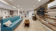 Located at 825 Pontiac Ave in Cranston, Rhode Island, this two-floor community facility offers a thoughtfully designed layout across a generous footprint. The upper floor serves as the main level, greeting visitors with a bright, open lobby anchored by a striking sputnik-style chandelier suspended from soaring ceilings. Light hardwood-style flooring, a bold blue accent wall, and a circular area rug create a polished, welcoming atmosphere. Workstations and seating areas flank the central space, while a beverage bar with granite countertops, dark cabinetry, and a mounted flat-screen TV provides a comfortable lounge amenity. A private office with vaulted ceilings, large windows, and open shelving offers a productive workspace bathed in natural light. The lower floor features a casual lounge area distinguished by a dramatic reclaimed wood accent wall with a mounted television, teal seating, and light vinyl plank flooring throughout. French doors connect to an adjacent dining or multipurpose area. The highlight of this level is a spacious Fitness Center, well-equipped with treadmills, ellipticals, a spin bike, cable machines, free weights, and a full dumbbell rack, all illuminated by recessed lighting and natural light from multiple windows. A utility and transition area with staircase access completes the lower level.