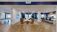 This single-story clubhouse in Panama City Beach, FL, showcases a cohesive coastal aesthetic defined by bold navy blue walls, crisp white wainscoting, and eye-catching herringbone-patterned wood flooring that flows throughout the entire space. The interior is organized into several distinct yet open zones, each thoughtfully designed for both comfort and functionality.

The main lounge area centers around a plush white sectional sofa accented with navy throw pillows, complemented by woven rattan armchairs and a wall-mounted flat-screen television. Oversized windows dressed in plaid drapery admit generous natural light and offer views of the surrounding landscape. A dramatic multi-arm pendant light anchors the seating arrangement.

Adjacent to the lounge, a dedicated game room features a full-size billiards table with a navy felt surface, a mounted television, and a separate seating area for casual socializing. A glass-walled private room provides a quieter retreat with its own distinctive botanical wallcovering.

The co-working area offers a long white countertop bar lined with navy upholstered stools, desktop computers, and pendant lighting in a black-and-gold finish. Nearby, a Starbucks-branded coffee station includes navy cabinetry, bold geometric tile, open shelving, and a white quartz serving counter, rounding out this well-appointed community gathering space.