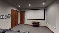 This single-level home theater room is thoughtfully designed to deliver a true cinema experience. The focal point of the space is a large, wall-mounted projection screen with a sleek black frame, positioned at the front of the room for optimal viewing from every seat. Opposite the screen, multiple rows of plush black leather power recliners are arranged in a tiered layout, each equipped with built-in cup holders and generous cushioning to maximize comfort during extended viewing sessions. The seating arrangement ensures clear sightlines to the screen from all positions.

The room is finished with neutral gray carpeting that enhances acoustics and adds to the theater ambiance. Recessed ceiling lights provide subtle, adjustable illumination, while a window fitted with horizontal blinds along one wall allows for effective light control. Warm wood baseboards and a solid wood entry door add a refined touch to the interior. A multi-tiered wooden media console near the screen offers convenient equipment storage.

Wall-mounted handrails assist with navigation between seating rows, and framed artwork, including a prominent nature photograph, decorates the rear wall, lending the space a polished, finished aesthetic. Overall, the room is well-appointed and purpose-built for immersive entertainment.