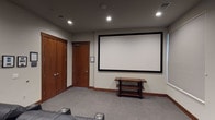 This single-level home theater room is thoughtfully designed to deliver a true cinema experience. The focal point of the space is a large, wall-mounted projection screen with a sleek black frame, positioned at the front of the room for optimal viewing from every seat. Opposite the screen, multiple rows of plush black leather power recliners are arranged in a tiered layout, each equipped with built-in cup holders and generous cushioning to maximize comfort during extended viewing sessions. The seating arrangement ensures clear sightlines to the screen from all positions.

The room is finished with neutral gray carpeting that enhances acoustics and adds to the theater ambiance. Recessed ceiling lights provide subtle, adjustable illumination, while a window fitted with horizontal blinds along one wall allows for effective light control. Warm wood baseboards and a solid wood entry door add a refined touch to the interior. A multi-tiered wooden media console near the screen offers convenient equipment storage.

Wall-mounted handrails assist with navigation between seating rows, and framed artwork, including a prominent nature photograph, decorates the rear wall, lending the space a polished, finished aesthetic. Overall, the room is well-appointed and purpose-built for immersive entertainment.