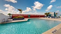 Located at 2810 University Square Drive in Tampa, Florida, this rooftop amenity deck offers a resort-style outdoor experience elevated above the surrounding cityscape. The centerpiece of the space is a generously sized swimming pool with crystal-clear water, complemented by a separate spa enclosure framed by a bold horizontal-slat privacy screen. Hexagonal-patterned stone pavers extend across the entire deck, providing a cohesive and durable surface throughout. Rows of blue lounge chairs line the poolside, offering ample space for sunbathing, while large spherical planters filled with mature palm trees add a tropical ambiance to the setting. Patches of manicured turf and lush tropical plantings introduce greenery and color against the clean, modern architecture. Shaded seating areas with umbrellas provide relief from the Florida sun. Tall pole lighting ensures the deck remains functional and inviting after dark. The perimeter wall opens up to sweeping panoramic views of the Tampa skyline and tree canopy, with open skies above creating a bright, airy atmosphere throughout. The overall design balances contemporary aesthetics with practical resort-style amenities, making it a standout outdoor gathering space.