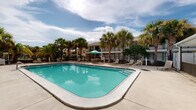 Located at 75 Nightingale Ln in Gulf Breeze, Florida, this outdoor pool area offers a classic Florida recreational experience in a well-maintained, open-air setting. The centerpiece of the space is a large rectangular swimming pool featuring clear turquoise water, gentle entry steps, and a surrounding concrete deck that provides ample room for lounging and circulation.

The pool deck is furnished with multiple lounge chairs and shaded seating areas complete with umbrella tables, offering guests comfortable spots to relax under the Florida sun. A dedicated pool house structure sits adjacent to the pool, providing practical amenities for guests. The entire area is enclosed by a black iron fence that ensures safety and privacy while maintaining an open, airy feel.

Tall, mature palm trees are planted throughout the perimeter, lending the space a tropical ambiance and natural shade. Lush greenery and additional trees line the fence line, creating a visually appealing and secluded atmosphere. The surrounding multi-story building with balconies is visible from the pool area, reinforcing the communal, resort-style character of the property. Under the expansive Gulf Coast sky, this pool area delivers a vibrant and inviting outdoor retreat suited for both relaxation and recreation.