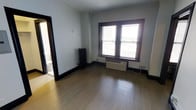 This single-floor space offers a straightforward and functional layout with several notable features. The main room is finished with light-toned hardwood flooring that runs throughout, lending a warm and clean aesthetic to the interior. Light-gray walls provide a neutral backdrop, while dark-stained wood trim along the baseboards, door frames, and window casing adds a classic contrast and architectural character.

Natural light enters generously through a pair of double-hung windows fitted with horizontal blinds, positioned along one wall and framed by the same dark wood trim. Below the windows sits a wall-mounted PTAC (packaged terminal air conditioning) unit, offering both heating and cooling capabilities. A traditional cast iron radiator is also present along the adjacent wall, indicating the space is equipped with radiant heating as well.

The room opens to an adjoining bathroom, visible through a doorway and featuring tiled flooring and standard fixtures. Crown molding runs along the ceiling, adding a subtle but refined architectural detail that elevates the overall finish of the space. The room is unfurnished, allowing for a clear appreciation of its dimensions and layout. Overall, the space presents a clean, utilitarian character with classic building details and practical climate control amenities.