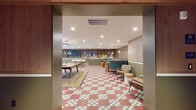 Located at 3850 Boston St in Baltimore's Canton neighborhood, this game room is a thoughtfully designed communal space that blends retro charm with modern comfort. The eye-catching red and white checkered tile flooring sets a playful tone throughout, anchored by warm wood-paneled walls painted in a two-tone scheme of natural oak and sage green. Recessed lighting keeps the space bright and welcoming. Gaming is central to the room's identity, with a foosball table, a shuffleboard table, and a pool table positioned atop area rugs for a relaxed, social atmosphere. A gallery wall featuring framed jerseys, pennants, and eclectic artwork adds personality and a retro sports-lounge aesthetic. Comfortable seating options include teal upholstered booth-style benches, mid-century modern lounge chairs, and drum-style side tables arranged around a round dining table. A wet bar with striking marbled countertops and a bold patterned cabinet front provides a functional entertaining hub. A wall-mounted TV offers additional entertainment options. Floor-to-ceiling glass doors open to an outdoor patio furnished with seating, bringing in natural light and extending the social space outdoors. The overall ambiance is energetic yet inviting, making this game room ideal for recreation and community gathering.