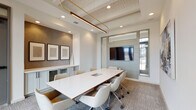 This single-floor conference room is a professionally designed meeting space that balances contemporary aesthetics with functional utility. At its center sits a large, sleek white conference table surrounded by ergonomic upholstered chairs on casters, comfortably accommodating groups for collaborative work or formal meetings. The room's coffered ceiling adds architectural depth, while a striking brass-and-wood linear pendant light serves as a refined focal point overhead, complemented by recessed lighting throughout.

One wall features a large flat-panel display mounted on a shiplap-paneled accent wall, framed by interior glass windows that allow views into adjacent spaces and maintain an open, connected feel. Opposite, a textured accent wall in a subtle geometric pattern hosts a trio of framed art pieces above a sage-green built-in credenza with mesh cabinet fronts, providing both storage and visual interest. An additional piece of abstract artwork adorns a side wall, adding a creative touch to the professional environment.

Natural light pours in through a set of tall, dark-framed glass exterior doors fitted with transom windows, offering views of the surrounding landscape. The patterned carpet adds warmth and texture underfoot, tying together the room's neutral palette of whites, grays, and warm wood tones. This space is ideally suited for business meetings, presentations, and collaborative sessions.