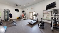 Located at 13621 W Glendale Ave in Glendale, Arizona, this fitness center is a dedicated exercise facility designed to meet a variety of workout needs. The space features clean white walls, dark rubber flooring, and recessed lighting complemented by a ceiling fan, creating a functional and well-lit environment for physical activity.

The room is outfitted with a comprehensive selection of cardio equipment, including multiple treadmills, a stationary bike, an elliptical trainer, and a rowing machine — providing options for a full range of cardiovascular workouts. For strength training, the space includes a cable machine, an adjustable weight bench, a rack of dumbbells, and various accessories such as medicine balls and stability balls.

Large wall-to-wall mirrors on one side of the room enhance the sense of openness and allow users to monitor their form during exercise. Multiple wall-mounted flat-screen televisions are positioned throughout for entertainment during workouts. A set of double glass doors leads to an outdoor area, allowing natural light to filter in and providing ventilation. The facility also includes a trash receptacle and safety signage, reflecting a practical and safety-conscious design. Overall, this fitness center delivers a well-equipped, purposeful space suited for daily exercise.