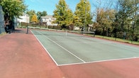 Blackbird Farms offers a well-maintained outdoor tennis court designed for year-round recreational use. The court features a classic hard surface finished in a green playing area with a deep red surround, clearly marked with crisp white boundary lines and fitted with a regulation-height net at center court. The surface provides consistent play conditions and is well-suited for both casual and competitive matches.

The court is fully enclosed by a tall black chain-link fence, which serves both as a ball-containment barrier and a defined perimeter for the facility. Access is provided through a dedicated gated entrance, allowing for easy and controlled entry. Tall light poles are positioned along the court, enabling play during evening hours and extending the usability of the space beyond daylight.

The surrounding environment enhances the outdoor experience, with mature trees framing the area and providing a natural backdrop. The lush greenery and open sky above create a pleasant, park-like atmosphere. The overall setting is clean and functional, reflecting a well-kept community amenity. Neighboring buildings are visible beyond the tree line, situating the court comfortably within the broader Blackbird Farms community.