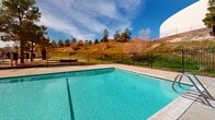 Located in Palmdale, California, this single-level outdoor recreational facility centers around a generously sized swimming pool with clear, blue-green water and a smooth tiled interior. The pool is complemented by a wide, well-maintained concrete deck that provides ample space for lounging and movement around the perimeter. Safety is well addressed through secure wrought iron fencing that fully encloses the pool area, complete with a gated entry and a life ring station for emergency use. A small pool house structure with a red tile roof sits adjacent to the pool, offering a classic Spanish-influenced architectural touch consistent with the surrounding buildings. The facility also features a spacious parking area enclosed by iron gates, framed by mature trees including palms and pines that add natural character to the grounds. The backdrop of the space includes a rolling hillside with scattered trees and a prominent white dome structure, giving the property a distinctive visual landmark. The open sky setting allows for abundant natural light throughout the day, enhancing the outdoor ambiance. Overall, this facility presents a well-equipped, functional outdoor amenity space suited for community recreational use in the high desert environment of the Antelope Valley.