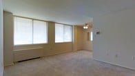 This single-floor residential space offers a clean, functional layout with a warm and understated aesthetic throughout. The living area is a standout feature, characterized by multiple large windows fitted with horizontal blinds that allow abundant natural light to fill the room, creating a bright and airy atmosphere. The walls are finished in a soft, neutral beige tone that complements the light-colored carpeting underfoot, contributing to a cohesive and calming interior palette. A ceiling fan with an integrated light fixture is centrally positioned in the main living area, providing both illumination and airflow. A baseboard heating unit along the wall ensures year-round comfort. The space transitions smoothly into an adjacent area, visible through an open doorway, which features an additional window that further enhances natural light flow. The overall layout is open and unobstructed, offering flexible use of the available space. White baseboards provide a crisp contrast against the neutral walls, adding a touch of refinement to the otherwise simple and practical design. The space presents a straightforward, move-in-ready environment well-suited for comfortable everyday living.