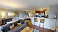 This single-floor studio offers a thoughtfully designed open-concept layout that combines the living, sleeping, and kitchen areas into one cohesive space. Warm hardwood flooring flows throughout, anchoring the room with a sense of continuity and warmth. The living area features a comfortable grey sofa with accent pillows in gold and cream tones, paired with a rustic wood coffee table that adds a natural touch. A circular area rug defines the sleeping zone, where a neatly styled bed with layered bedding sits alongside a compact nightstand and lamp. The kitchen is equipped with stainless steel appliances, including a refrigerator, microwave, and range, all set beneath rich, warm-toned cabinetry. A granite-topped breakfast bar with modern industrial-style bar stools serves as a natural divider between the kitchen and living space, offering both functionality and visual separation. Accent walls in a soft blue-grey hue add depth and color contrast to the neutral palette. A short hallway visible in the background suggests access to a bathroom, maintaining a practical flow throughout the unit. The overall ambiance is modern, cozy, and efficiently organized, making excellent use of the available space.