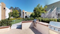 Located at 6675 Jafte Ct in San Diego, this well-appointed outdoor courtyard serves as a central communal amenity within a multi-building residential community. The courtyard is thoughtfully designed with a paver-tiled surface that extends throughout, providing a clean and durable foundation for outdoor activity and gathering.

A standout feature of the space is the built-in outdoor BBQ grill station, constructed with a stucco-finished counter and equipped with a stainless steel gas grill. The station is designed for convenience and social use, making it ideal for outdoor cooking and entertaining. Flanking the grill area are low stucco walls with warm-toned wood-cap seating ledges that add both function and aesthetic appeal.

Lush, mature landscaping surrounds the courtyard, featuring a variety of shrubs, ornamental grasses, flowering plants, and established trees that provide shade and a vibrant natural atmosphere. A concrete walkway cuts through manicured lawn areas, connecting the courtyard to the surrounding buildings, which feature stucco exteriors, balconies with metal railings, and exterior staircases.

A laundry center is conveniently accessible directly from the courtyard, clearly marked with signage and a secure entry door. The space benefits from abundant natural light under San Diego's clear blue skies, creating a bright and welcoming outdoor environment year-round.