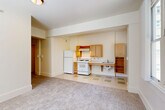 Located at 706 9th Ave in Seattle, WA, this efficiently designed studio unit offers a thoughtfully organized single-floor layout. The combined living, dining, and kitchen area forms the heart of the space, where light-toned carpet flows seamlessly into a kitchen alcove finished with easy-care vinyl tile in a green and cream checkered pattern. The kitchen is outfitted with light maple cabinetry, a gas range, refrigerator, and a wall-mounted counter with an integrated sink — a layout that maximizes functionality within a compact footprint. Large double-hung windows flood the main living area with natural light and frame views of surrounding trees and the Seattle urban streetscape, while a classic cast-iron radiator beneath the windows provides reliable warmth. The crisp white walls and neutral palette throughout create a clean, open ambiance. The accessible bathroom is equipped with stainless steel grab bars, reflecting a thoughtful, ADA-conscious design. A dedicated walk-in closet fitted with wire shelving and a hanging rod offers organized storage. An intercom system at the entry adds a practical security feature. The overall aesthetic is simple, bright, and functional — well-suited for efficient urban living in the heart of Seattle.