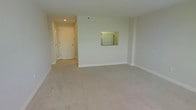 This single-floor residence offers a straightforward and functional layout designed with practicality in mind. The living area is generously proportioned, featuring wall-to-wall carpeting in a neutral beige tone that adds warmth and comfort underfoot. The walls are painted in a clean, light off-white finish, contributing to a bright and airy atmosphere throughout the space.

A distinctive architectural detail is the pass-through window cut into the wall between the living area and the kitchen, allowing visual connectivity between the two spaces and making the layout feel more open and communicative. Through the pass-through, a refrigerator is visible, indicating a well-equipped kitchen area beyond.

The entryway is clearly defined, featuring a solid white door with clean trim and recessed lighting overhead that provides focused illumination at the entrance. The hallway leading from the entry is neatly finished with consistent flooring and white baseboards that run throughout, tying the spaces together cohesively.

Overall, the space presents a clean, move-in-ready environment with a neutral color palette that provides a versatile canvas for a variety of interior styles. The understated design and efficient use of space make it a practical and adaptable living environment.