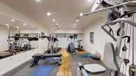 Located at 1636 N Logan St in Denver's Capitol Hill neighborhood, this dedicated fitness center offers a thoughtfully organized workout space designed to support a full range of exercise routines. The room features a combination of hardwood flooring and interlocking rubber mat zones, providing both durability and safety for various types of training. Recessed lighting throughout the ceiling ensures the space is well-lit and functional at any hour.

The cardio section is equipped with a treadmill, an elliptical trainer, and a stationary spin bike, offering ample options for aerobic conditioning. Strength training is well represented with a multi-functional cable and weight machine, a power tower for pull-ups and dips, an adjustable weight bench, a dumbbell rack, and a rowing machine. Stability and flexibility accessories, including a stability ball and a balance dome, are also on hand for functional fitness training.

A large wall-mounted mirror spans one side of the room, visually expanding the space and allowing users to monitor their form during workouts. White walls and clean finishes contribute to a bright, motivating atmosphere. The fitness room is accessed through a dedicated door clearly labeled "Fitness," reinforcing its purpose as a professional-grade workout facility.