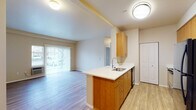 This single-floor residence is designed with a practical, open layout that connects the kitchen and living areas seamlessly. The kitchen is equipped with warm-toned wood cabinetry, white countertops, a peninsula with an undermount sink, black appliances including a refrigerator, electric range, microwave, and dishwasher, along with a pantry accessed via bifold doors. Light wood-style flooring runs throughout the main living and kitchen spaces, contributing to a bright, cohesive aesthetic. The living area opens to a sliding glass door fitted with vertical blinds, providing outdoor access and natural light. In-unit laundry is conveniently located in a dedicated closet adjacent to the main living space. The bedrooms are carpeted and feature baseboard heating, windows with horizontal blinds offering outdoor views, and ample wall space. The bathroom includes a wood-toned vanity with a white countertop, a large mirror, and a full bathtub and shower combination. Throughout the unit, neutral beige walls and consistent finishes create a clean, functional living environment.