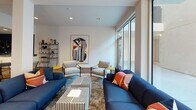 The Club Room at 417 Centinela Ave in Inglewood, California is a thoughtfully designed single-floor communal amenity space that blends contemporary style with functional versatility. The open-plan layout is anchored by rich hardwood flooring and a bold deep navy accent wall that creates a striking visual contrast against the crisp white ceilings and walls throughout.

The lounge area features a pair of large navy blue sofas accented with vibrant orange and coral throw pillows, arranged around a bold black-and-white striped coffee table set. Flanking wall sconces and large floor-to-ceiling windows invite an abundance of natural light, enhancing the warm, inviting ambiance. Curated abstract artwork adorns the walls, adding a gallery-like quality to the space.

The dining and gathering zone offers a long high-top communal table with sleek grey upholstered chairs, complemented by a separate bistro-style round table with wire-frame seating — ideal for both casual meetups and collaborative sessions. A built-in bench with decorative mosaic tile detailing runs along one wall, providing additional flexible seating.

The kitchenette features white cabinetry, a sink, and stainless steel appliances, accented by a dramatic living plant wall display against the navy tile backsplash. Open shelving units throughout the space display curated décor objects, completing this polished and functional club room.