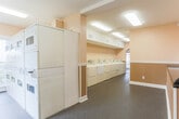 Located at 728 Frawley Rd in East Ridge, Tennessee, this single-floor community amenity facility is designed to serve residents with a range of practical and recreational spaces. The laundry room is the centerpiece of the facility, featuring a generous row of full-sized top-loading washers and dryers lined along the wall, all finished in white and equipped with modern controls. A separate alcove houses stacked washer-dryer units alongside a window that brings in natural light, with a seating area nearby for convenience. The flooring throughout the laundry area consists of durable speckled vinyl, well-suited for high-traffic use. Adjacent to the laundry room is a spacious lounge and waiting area finished with neutral beige walls accented by two-tone wainscoting and plush grey carpet. This area features a built-in countertop ledge, a cushioned chair, and a glass-paned door that connects to the exterior, allowing additional natural light to filter in. Overhead fluorescent lighting ensures consistent illumination throughout. Safety equipment, including a fire extinguisher and signage, is prominently installed. A glimpse into an adjoining fitness area reveals exercise equipment, rounding out the facility's amenities. The overall design is clean, functional, and community-oriented.