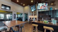 This clubhouse is a thoughtfully designed community hub that seamlessly blends social, recreational, and fitness amenities under one roof. The space is anchored by soaring ceilings that lend an open, airy atmosphere throughout. Warm hardwood-style flooring flows across the entire layout, unifying the distinct zones within the space.

The lounge area offers a comfortable gathering spot, furnished with a generous dark leather sectional sofa, colorful accent pillows, and a soft ottoman. Large windows dressed with sheer curtains and blinds flood this area with natural light while providing views of the surrounding exterior. Tall ceilings with exposed beam detailing add architectural character to this relaxed seating zone.

Adjacent to the lounge is a bar-style kitchen and social area featuring dark cabinetry, granite-style countertops, and rustic wooden bar stools. Multiple flat-screen televisions are mounted overhead, making this a lively entertainment hub. Eclectic wall décor and colorful accents contribute to the space's vibrant, welcoming personality.

The communal area offers tall wooden high-top tables paired with industrial-style stools beneath a statement pendant light fixture, ideal for casual gatherings or collaborative use.

Directly accessible from the main area, the fitness center is visible through glass partitions and is equipped with cardio machines, weight equipment, and wall-mounted televisions, providing a fully functional workout space within the clubhouse.
