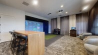 Located at 9875 W 58th Ave in Arvada, Colorado, this thoughtfully designed golf simulator room offers a premium indoor sports and entertainment experience within a well-appointed single-level layout. The centerpiece of the space is a Full Swing golf simulator, complete with a large projection screen, a dedicated hitting mat, and a rack stocked with golf clubs, enabling players to access a comprehensive games library that includes golf courses, football, baseball, and more. A ceiling-mounted projector delivers a vivid, immersive display against a dark-framed impact screen framed by floor-to-ceiling curtains. Adjacent to the simulator area, a natural wood high-top communal table with bar-height stools provides seating for spectators and social gatherings. A media console with a wall-mounted flat-screen TV and under-counter refrigerator offers added convenience for lounging guests. Golf-themed framed artwork accents the neutral gray walls, contributing to the space's polished aesthetic. On the opposite end of the room, a comfortable seating nook features plush curved lounge chairs positioned in front of expansive windows dressed with layered draping curtains, allowing natural light to filter in. Patterned carpet throughout ties the room together, creating a cohesive and inviting atmosphere ideal for recreation and entertainment.