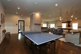 This single-floor space is thoughtfully designed as a multifunctional entertainment and recreation venue, combining several distinct zones within an open, flowing layout. Rich hardwood flooring runs continuously throughout, anchoring the modern farmhouse aesthetic that defines the space.

The central lounge area serves as the heart of the space, furnished with a generous sectional sofa, accent chairs, and a woven area rug. A chevron-patterned accent wall clad in light wood paneling provides a striking backdrop for a wall-mounted flat-screen television, while a dramatic cluster of globe pendant lights with exposed black cording hangs overhead, creating an artistic focal point.

Adjacent to the lounge, a dedicated game room houses a dark-stained pool table beneath a rustic recessed wood chandelier fitted with Edison bulbs. A cue rack and floating wood shelves line the wainscoted walls, and natural light filters in through multiple windows. A ping-pong table occupies a separate zone, accessible through a wide barn-style doorway framed in reclaimed wood.

The bar area features a sleek dark-paneled counter with high-gloss black tile, white upper shelving, and a herringbone tile backsplash. Lantern-style pendant lights illuminate the space, which also includes comfortable lounge seating and a small café-style table, creating an inviting gathering spot throughout.