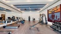 This fitness center at 1300 Front Ave in Columbus, Georgia, offers a comprehensive workout facility designed to meet a wide range of exercise needs. The space is organized across a single floor and is divided into distinct workout zones, each tailored to a specific type of training. The cardio area features multiple treadmills, ellipticals, and stationary bikes, all positioned near large floor-to-ceiling windows that flood the space with natural light and provide views of the outdoors. A full-length wall mirror enhances the sense of openness in this section. The strength training zone is well-appointed with a multi-station cable machine, a Smith machine with an adjustable bench, and a fully stocked dumbbell rack ranging across a variety of weights. The ceiling design is a standout architectural feature, showcasing a tiered tray ceiling accented with a bold blue inset panel, recessed lighting, and dual ceiling fans that maintain comfortable airflow throughout. Wood-look flooring in the cardio area transitions to a durable tile surface in the free weights section, clearly delineating functional zones. The overall ambiance is modern and motivating, combining clean white walls, vibrant accent artwork, and an abundance of natural light to create an inviting environment for fitness enthusiasts.