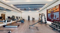 This fitness center at 1300 Front Ave in Columbus, Georgia, offers a comprehensive workout facility designed to meet a wide range of exercise needs. The space is organized across a single floor and is divided into distinct workout zones, each tailored to a specific type of training. The cardio area features multiple treadmills, ellipticals, and stationary bikes, all positioned near large floor-to-ceiling windows that flood the space with natural light and provide views of the outdoors. A full-length wall mirror enhances the sense of openness in this section. The strength training zone is well-appointed with a multi-station cable machine, a Smith machine with an adjustable bench, and a fully stocked dumbbell rack ranging across a variety of weights. The ceiling design is a standout architectural feature, showcasing a tiered tray ceiling accented with a bold blue inset panel, recessed lighting, and dual ceiling fans that maintain comfortable airflow throughout. Wood-look flooring in the cardio area transitions to a durable tile surface in the free weights section, clearly delineating functional zones. The overall ambiance is modern and motivating, combining clean white walls, vibrant accent artwork, and an abundance of natural light to create an inviting environment for fitness enthusiasts.