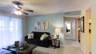 This single-story space presents a welcoming living area distinguished by its soft teal accent wall, which serves as a calming focal point against the surrounding neutral-toned walls. The room features wall-to-wall carpeting in a light beige hue, contributing to a warm and comfortable atmosphere throughout. A ceiling fan is mounted overhead on a textured popcorn ceiling, providing both airflow and a practical lighting source for the space.

The living area is well-proportioned, accommodating a dark upholstered sofa accented with decorative coastal-themed pillows, complemented by a wooden side table and a table lamp that adds a soft, ambient glow. Tropical palm tree artwork adorns the teal accent wall, reinforcing a relaxed, coastal aesthetic. A low-profile coffee table anchors the seating arrangement, resting on a textured area rug.

A wide doorway from the living area opens into a hallway, offering a glimpse of additional rooms beyond, including what appears to be a bedroom and other living spaces. The hallway is well-lit with overhead lighting, enhancing the overall sense of openness and connectivity throughout the single-floor layout.