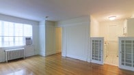 This single-floor space showcases a blend of classic architectural charm and functional design. The interiors are defined by elegant white wainscoting and raised panel wall detailing that add a sense of craftsmanship and traditional character to each room. Warm-toned hardwood floors run throughout, reflecting natural light and contributing to an inviting, cohesive ambiance.

The main living area is generously proportioned, with smooth white ceilings and clean-lined moldings that frame the space with understated sophistication. A window unit provides climate control, while a window along the wall allows natural light to filter in, brightening the room. A charming glass-paned cabinet or built-in display unit is tucked into one corner, offering both storage and a decorative focal point with its classic divided-light door design.

Doorways with paneled surrounds lead to additional rooms, maintaining the consistent architectural language found throughout the space. The neutral white and off-white palette creates a versatile backdrop well-suited to a variety of interior styles. Overall, the space presents a well-preserved, character-rich environment that balances traditional detailing with a clean, airy aesthetic.