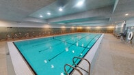 Located in Chicago's West Loop neighborhood, this single-level indoor aquatic facility at 1515 W Monroe St offers a well-appointed environment designed for both fitness and relaxation. The centerpiece is a generously sized, multi-lane lap pool with clearly marked swimming lanes and vivid turquoise water, accessible via stainless steel entry rails. Recessed overhead lighting illuminates the pool evenly, reflecting off the water's surface and creating a bright, functional atmosphere.

Encircling the pool, the walls are clad in a distinctive decorative mosaic tile mural depicting marine life — including colorful fish, coral, and sea flora — rendered in earth tones and cool blues. This artistic detail extends throughout the space, giving the facility a unique, visually engaging character. A separate wading or shallow-end area is positioned at one end of the pool, accompanied by its own set of entry steps.

Adjacent to the pool, a dedicated lounge area features cushioned wooden armchairs arranged along the mosaic-tiled walls, offering comfortable seating for relaxation before or after swimming. The flooring throughout the surrounding deck is composed of small, textured tiles, providing both a practical and cohesive aesthetic. The overall ambiance is calm and well-maintained, suited for regular aquatic use.