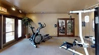 This fitness center is a thoughtfully designed, single-floor exercise facility offering a comprehensive range of workout equipment in a welcoming environment. The space is outfitted with multiple treadmills and elliptical machines for cardio training, alongside a multi-station Life Fitness cable and weight machine, a leg press, and a free weight bench, providing users with a full-body workout experience.

The room features floor-to-ceiling mirrors along one wall, which visually expand the space and allow users to monitor their form during exercise. Track lighting runs across the ceiling, providing bright, even illumination throughout the room, complemented by recessed lighting. The textured ceiling adds a subtle architectural detail to the warm, neutral-toned walls.

Large French doors with glass panels line the exterior wall, inviting generous natural light into the space and offering views of an outdoor area, creating an open and airy atmosphere. Decorative potted palm plants are positioned throughout the room, adding a touch of greenery and enhancing the overall ambiance.

Carpeted flooring provides a comfortable surface underfoot, and posted fitness center rules and instructional charts on the walls reflect a focus on safety and proper equipment use. The space is well-organized, clean, and designed to support a productive and motivating workout environment.