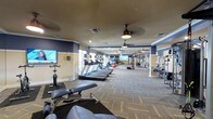 This fitness center offers a comprehensive and thoughtfully designed workout environment across a single, expansive floor. The space is anchored by a long row of treadmills and elliptical machines positioned along the windowed wall, allowing natural light to filter in and create an energizing atmosphere during cardio sessions. Complementing the cardio zone, a dedicated strength training area features a robust selection of weight machines, free weights, and benches, catering to a wide range of exercise routines. The patterned, low-pile carpeting adds a refined aesthetic while providing a comfortable underfoot surface throughout. Overhead, a series of ceiling fans with pendant lighting ensures consistent air circulation and a well-lit environment. Flat-screen TVs are mounted on the walls, offering entertainment options to keep workouts engaging. The walls are finished in a warm two-tone palette of soft gold and deep navy blue, trimmed with classic crown molding, lending the space an upscale feel. Stability balls and additional accessories are neatly arranged throughout, rounding out the functional offerings of this well-appointed fitness facility.