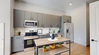 Located at 2100 Abbey Ave in Cleveland's Ohio City neighborhood, this single-floor unit offers a thoughtfully designed layout that maximizes its compact footprint. The interior features a clean, modern aesthetic defined by light wood-style flooring, crisp white walls, and black hardware accents throughout.

The open-concept kitchen is a standout feature, equipped with stainless steel appliances including a range, microwave, and refrigerator. Flat-front gray cabinetry extends to the ceiling, complemented by a subway tile backsplash and granite-style countertops. A central island provides additional prep and dining space.

The living area benefits from notable ceiling height, creating an airy, spacious feel. A large window fitted with horizontal blinds allows natural light to filter in while offering views of the surrounding greenery. The dining area is positioned near this window, enhancing the bright atmosphere.

The bedroom features soaring ceilings, soft carpet flooring, and a frosted glass sidelight adjacent to the door, allowing light without sacrificing privacy. The bathroom is finished with wood-look tile flooring, a gray vanity with a white countertop, and a large mirror. A hallway connects the spaces efficiently, completing this well-appointed unit.